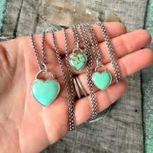 Three heart-shaped turquoise pendants on silver chains held in a hand against a textured fabric background.