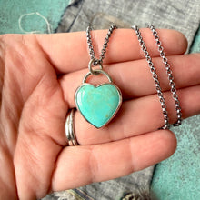 Turquoise heart-shaped pendant on a silver chain held in a hand against a textured background.
