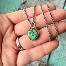 Turquoise heart-shaped pendant on a silver chain held in a hand against a textured background.